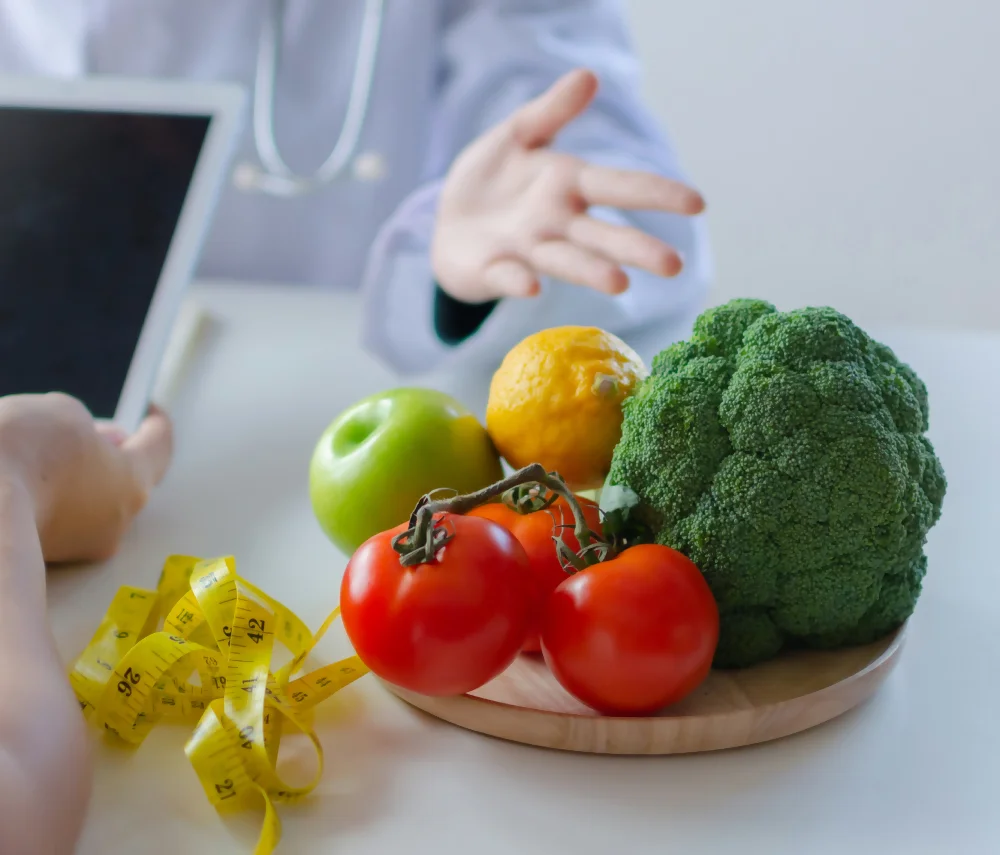 In the nutrition consultation, the nutritionist points out a plate of fruits and vegetables while a measuring tape is seen on the table.