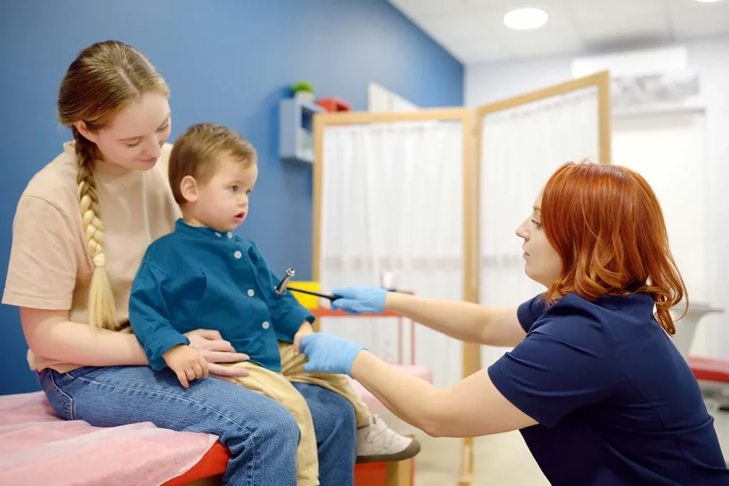 Neurólogo pediatra en consulta, revisando los reflejos de un niño pequeño mientras su madre lo sostiene.