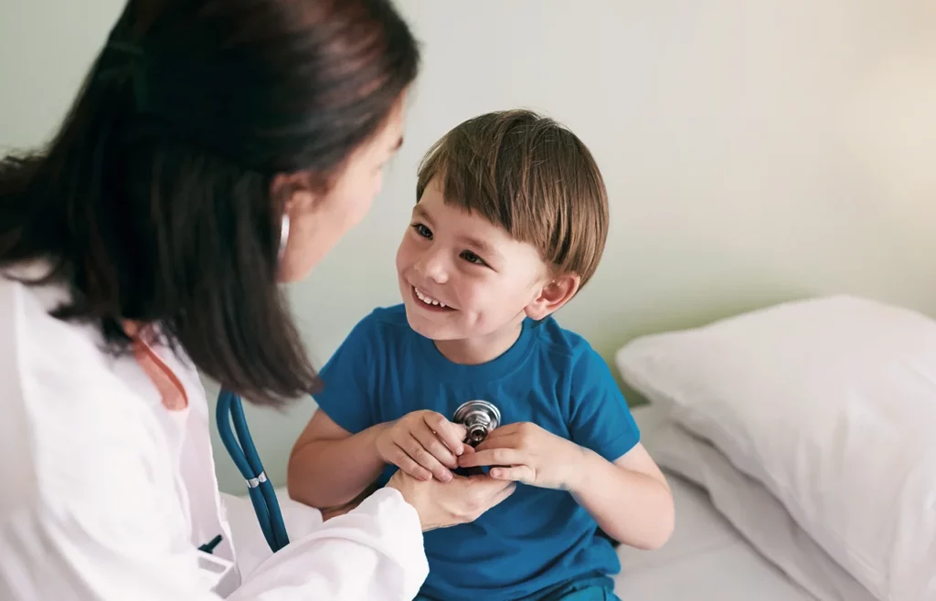 Niño, paciente de cardiocirugía pediátrica, sonriéndole a su doctora mientras esta le hace un chequeo de los latidos de su corazón.